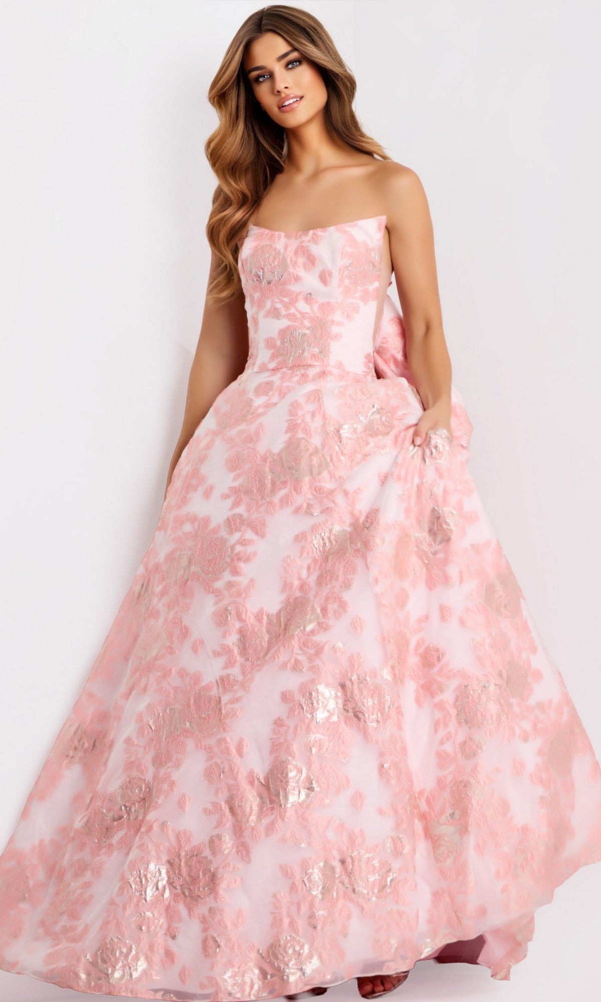 A woman poses elegantly against a plain light background wearing the Jovani 47702 long prom dress—a strapless jacquard ball gown with metallic pink floral patterns.